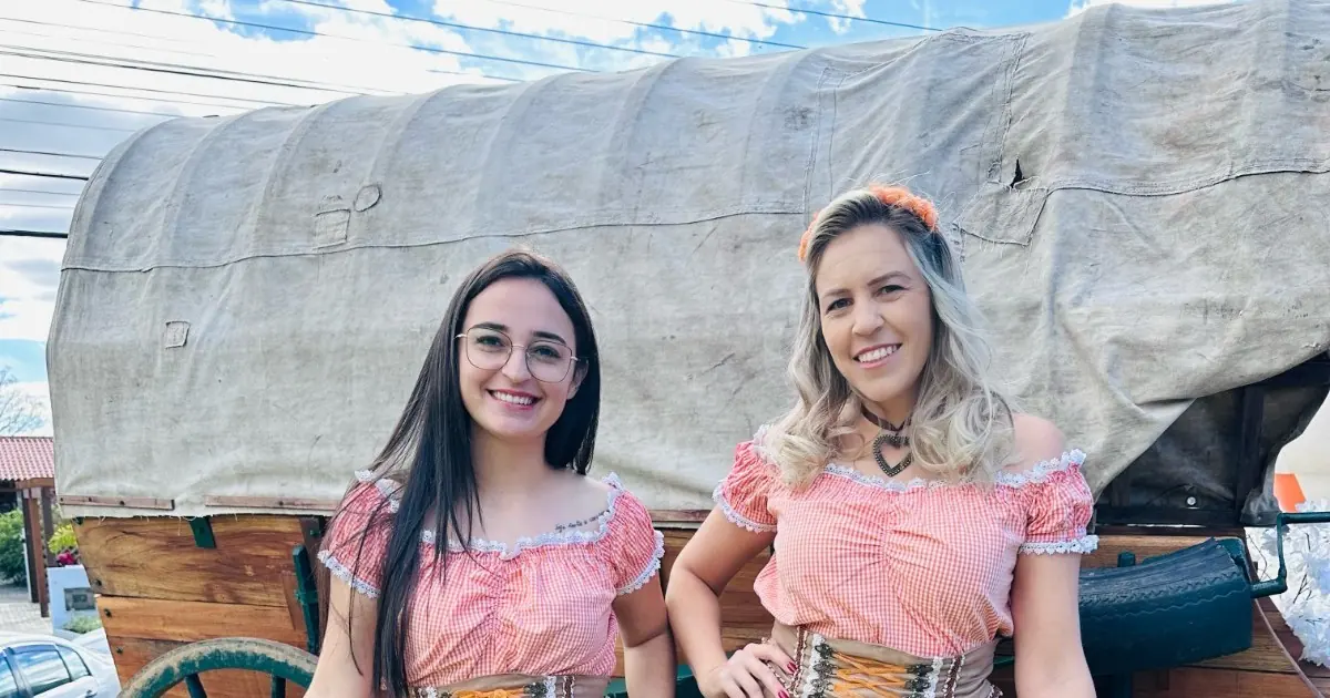 Duas mulheres com trajes típicos sorriem diante de uma carroça coberta da decoração