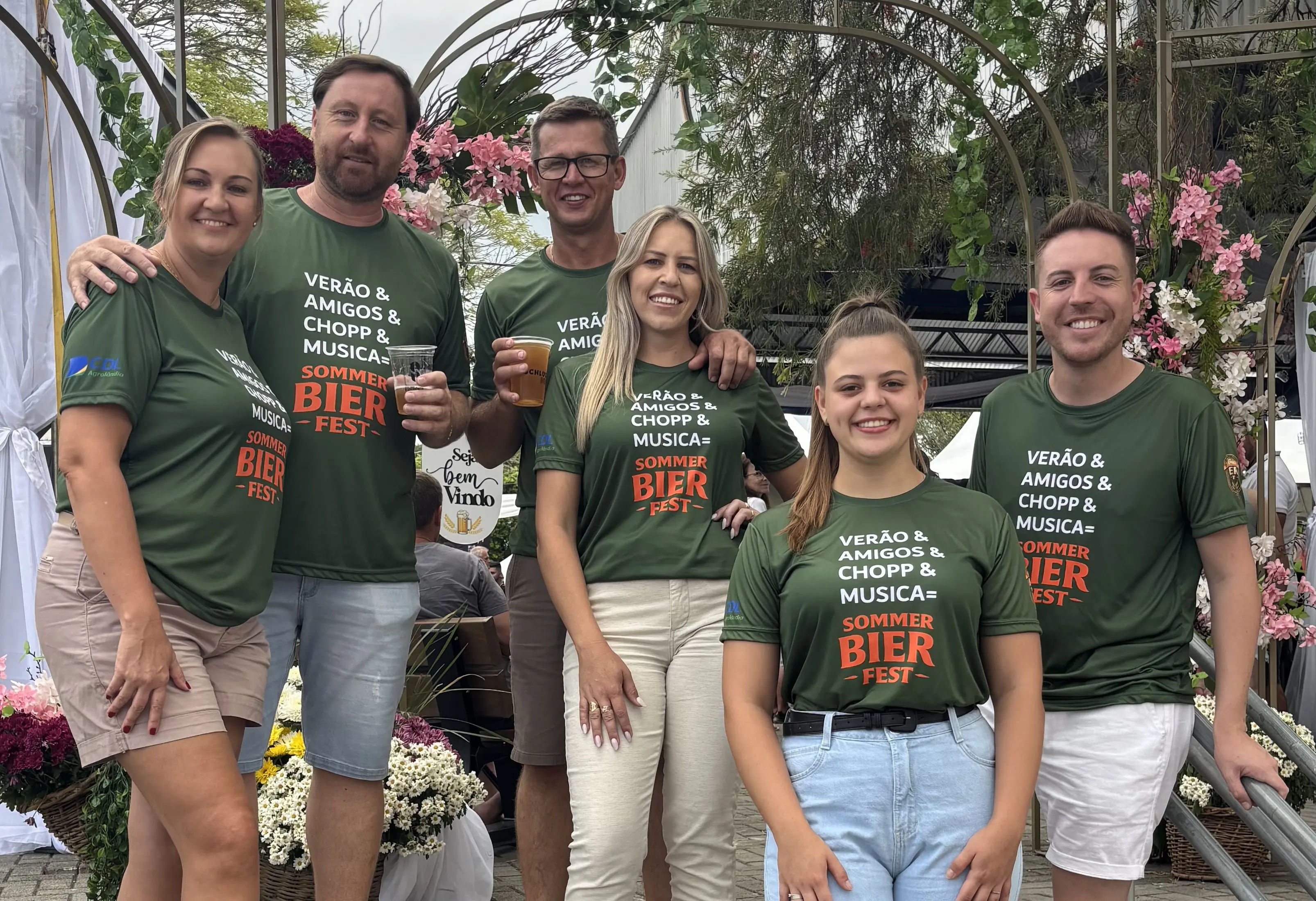 Equipe do Sommer Bier Fest com camisetas verdes do festival na escadaria da praça, sorrindo