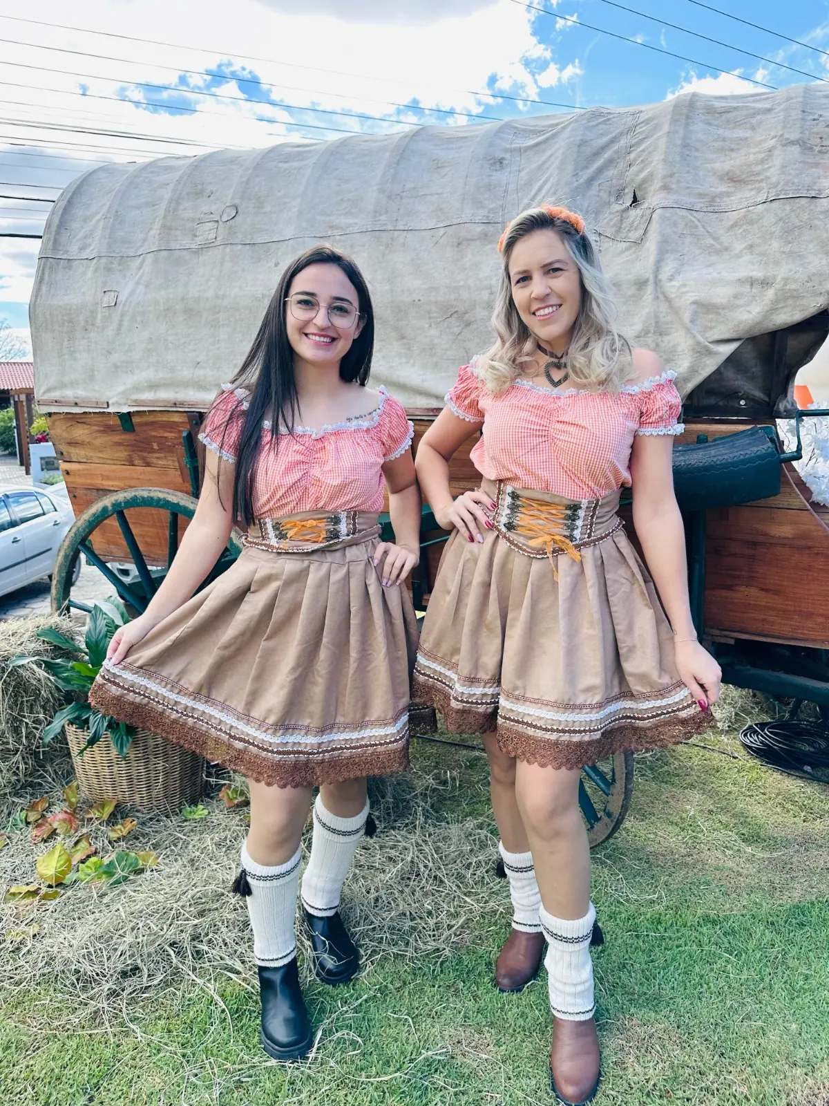 Duas mulheres com trajes típicos alemães ao lado de uma carroça coberta na decoração do festival