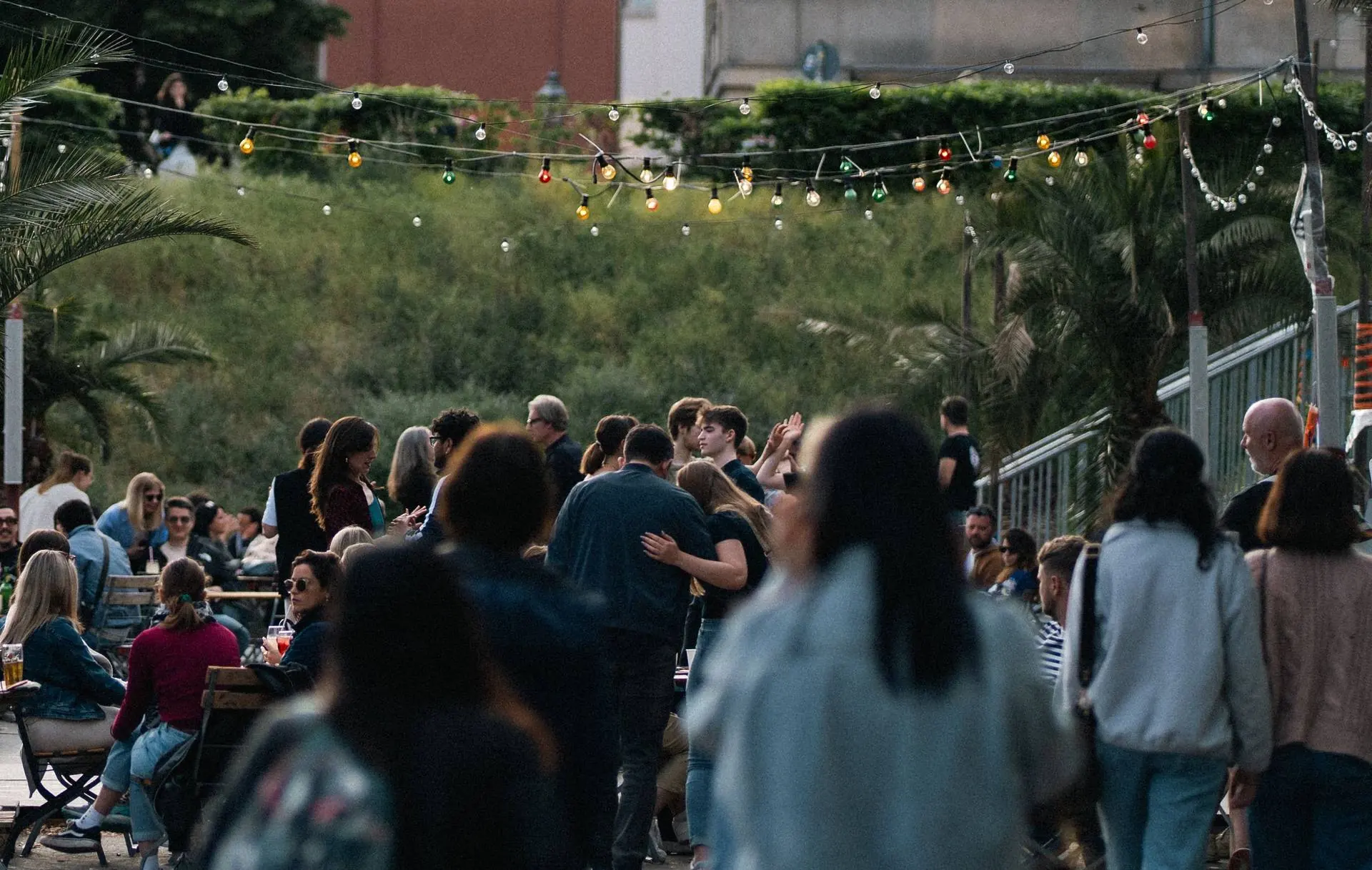 Festa de verão ao ar livre, com varal de lâmpadas coloridas e pessoas reunidas às mesas ao entardecer