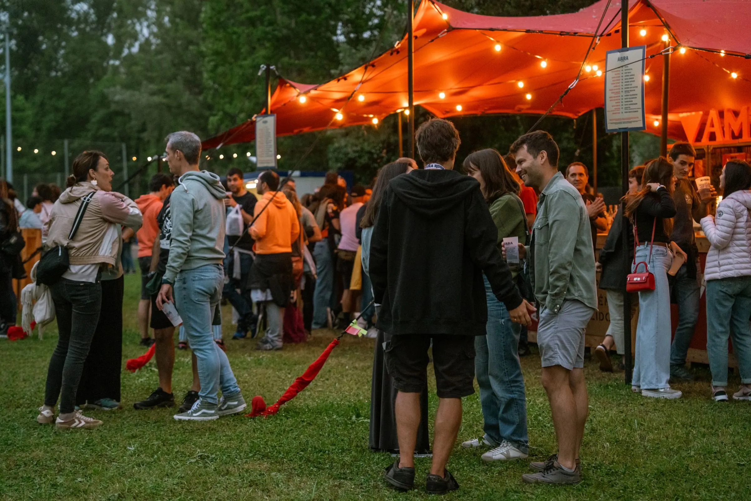 Grupo de adultos conversando no gramado de um festival ao ar livre, com tenda iluminada ao fundo