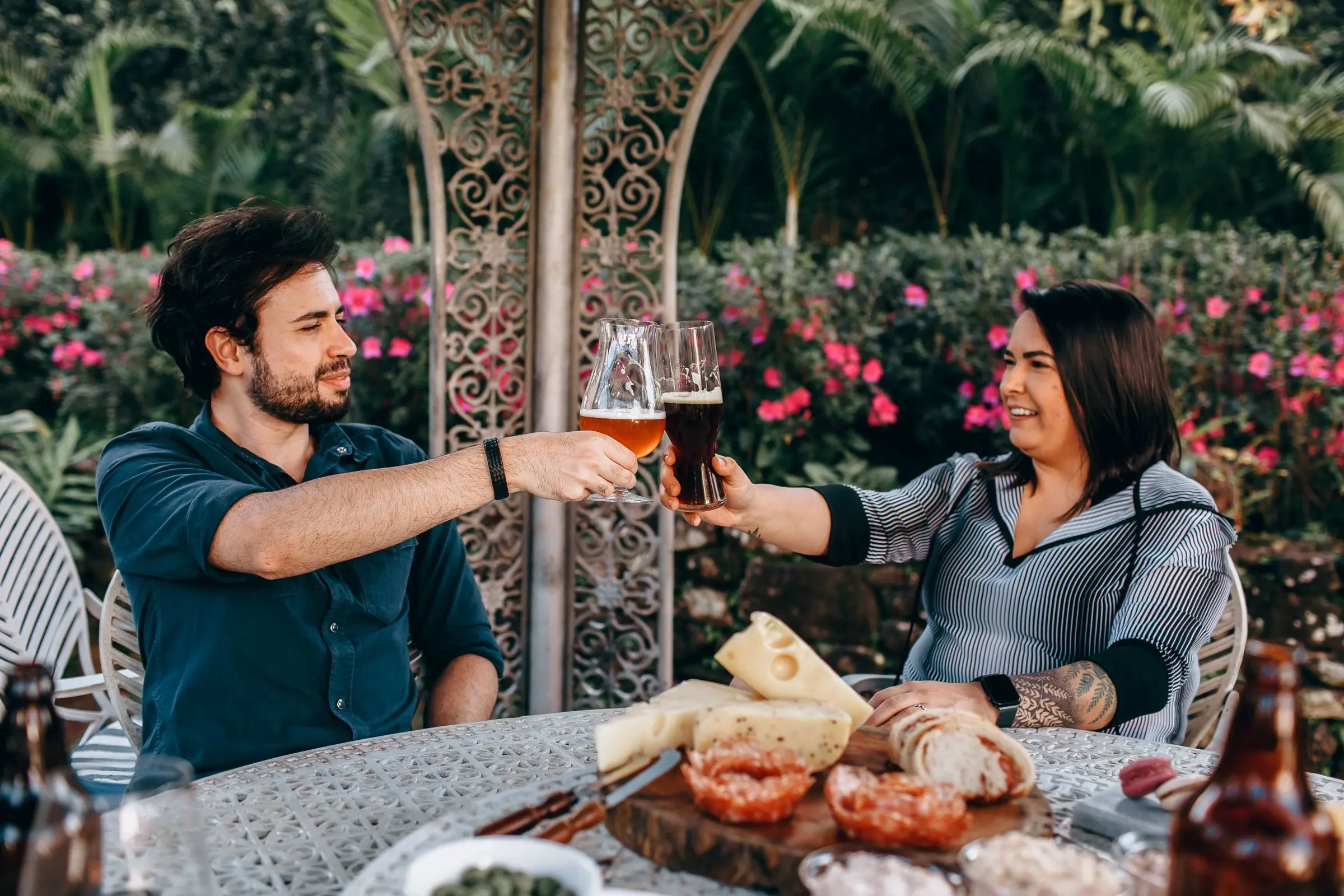 Homem e mulher brindando com copos de cerveja em uma mesa ao ar livre, cercados de plantas floridas